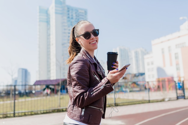 Outsdie portrait of charming smiling pretty woman with dark hair dressed leather jacket and black sunglasses listening to music in earphones