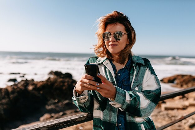 Outside portrait of attractive woman with curly short hairstyle wearing striped shirt using smartphone on shore of ocean with rocks