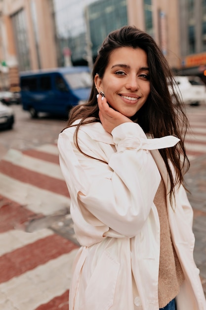 Outside portrait of attractive woman with dark hair wearing white coat walking on the street with charming smile