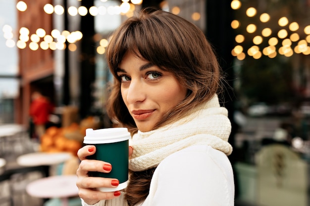 outside portrait of charming pretty woman with dark hair wearing white knitted sweater drinking coffee on lights background