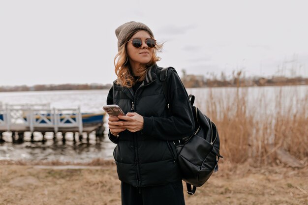 Outside portrait of stylish girl in sunglasses is using smartphone while standing against lake Girl is walking on the beach I'm warm sunny day