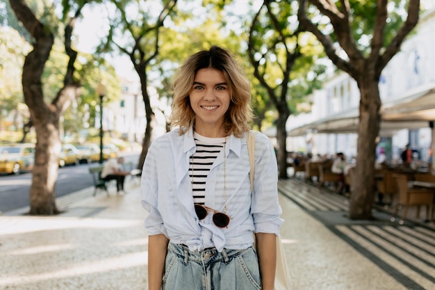 Outside portrait of young smiling woman with wavy hair wearing striped tshirt and blue shirt is posing at camera with happy smile against city square with green exotic trees