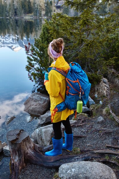 여성 여행자의 외부 샷은 파노라마 산악 호수를 즐기고, 산책 후 휴식 중에 따뜻한 차를 마시고, 큰 배낭을 들고, 휴가 여행을 가짐