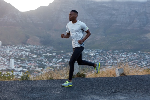 Outside shot of sporty dark skinned man in casual clothing, runs quickly, covers long distance, models over mountain landscape, wants to reach finish first. Athletic ethnic male poses outdoor