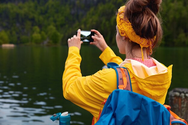 Outside shot of woman traveler makes photo of beautiful landscape on smart phone device, admires calm lake