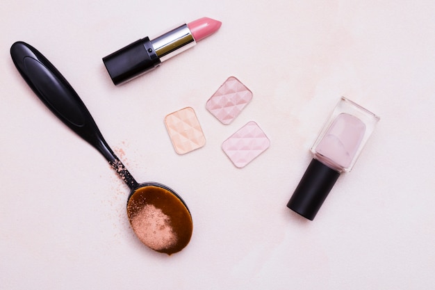 Oval black brush; compact powder; lipstick; nail varnish on pink background