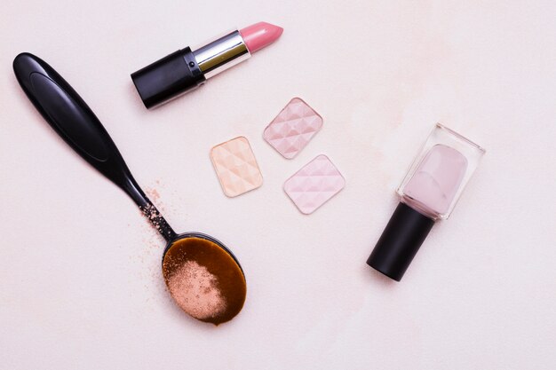 Oval black brush; compact powder; lipstick; nail varnish on pink background