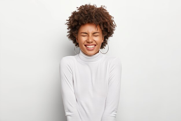 Overemotive Afro American lady laughs out positively, keeps eyes shut, smiles at funny story, expresses good emotions, wears white clothing, isolated, has curly haircut. People and emotions.