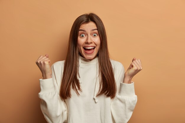 Overemotive dark haired woman has very happy look, clenches fists with joy, rejoices victory, opens mouth, has funny expression, wears casual hoodie