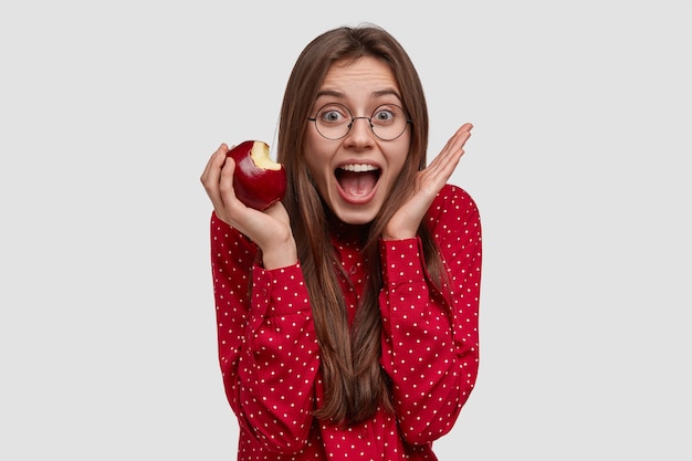 Overemotive happy lady raises hands near face, opens mouth widely, eats tasty apple, wears transparent glasses