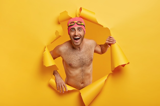 Overemotive man swimmer poses shirtless, wears swimhat and goggles, poses in torn paper hole, laughs happily