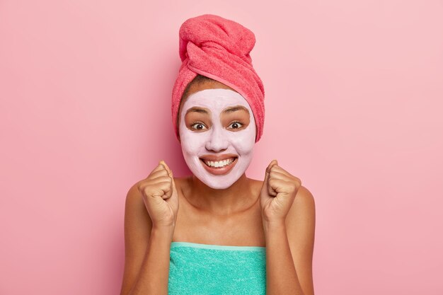 Overemotive woman supples facial skin, raises clenched fists, looks positively at camera, wears wrapped towel on head and body