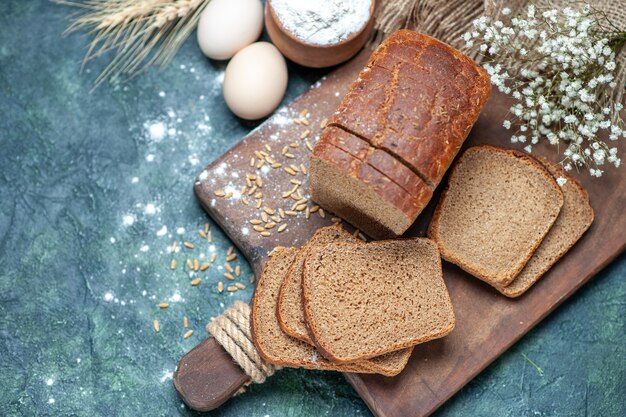 Overhead of dietary black bread wheats on wooden board spikes flower eggs flour in bowl on blue background