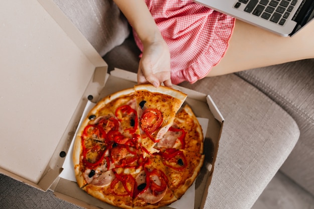 Overhead portrait of girl in checkered shorts eating pizza on sofa. Relaxed white woman enjoying snacks while working with laptop.