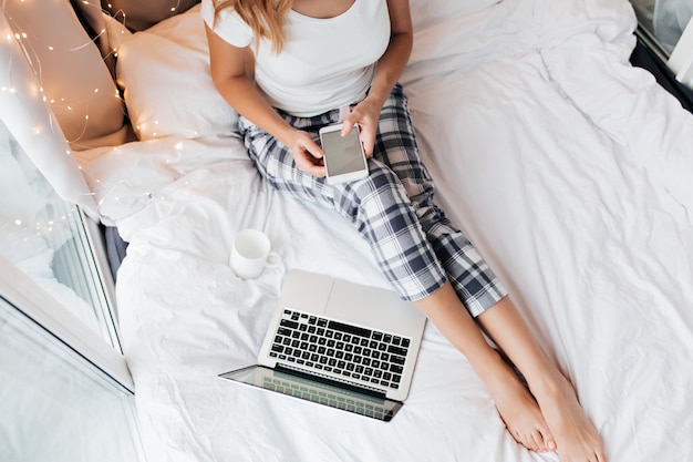 Overhead portrait of girl with smartphone sitting on white sheet. Indoor shot of female student doing homework with laptop in bed.