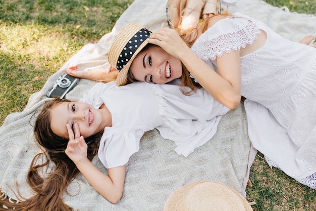 Накладные портрет расслабляющей девушки, лежа на одеяле и улыбаясь. Возбужденная молодая женщина отдыхает на траве, играя с радостной дочерью.