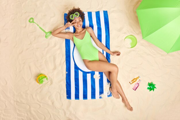 Снимок сверху веселой кудрявой женщины в маске для подводного плавания и зеленом купальнике делает мирный жест над позами глаз на полосатом полотенце на пляже с различными предметами вокруг концепции летнего времени