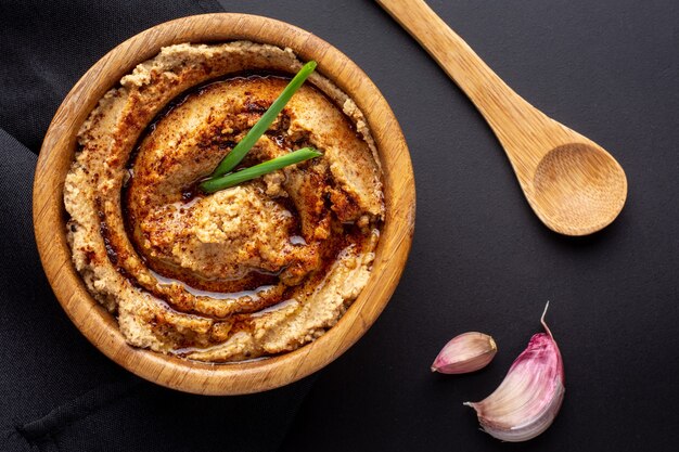 Overhead shot of  hummus in a wooden bowl with a wooden spoon and garlic pieces on black table