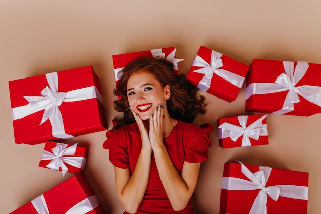 Overhead shot of lovely ginger woman enjoying birthday. Good-humoured red-haired female model lying on the floor beside presents.