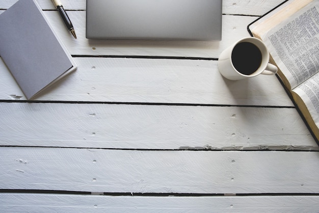 Overhead shot of white wooden surface with laptop, bible, coffee and a notepad with a pen on top