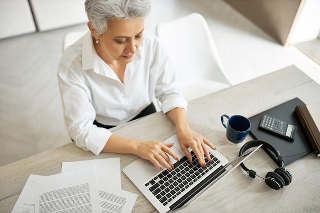 Free photo overhead top view of successful experienced mature female translator or copywriter in formal clothing sitting at office desk with mug, papers, headphones and laptop, typing text