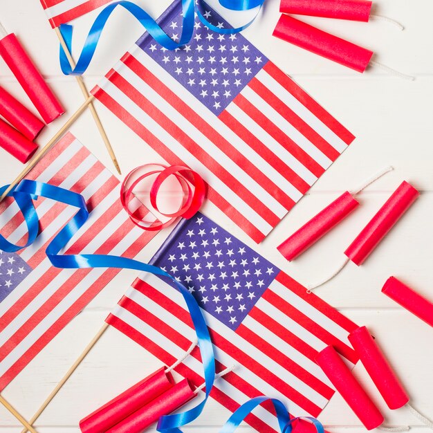 An overhead view of american flags with ribbons and firecracker on white desk