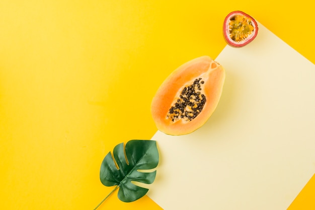 An overhead view of artificial leaf; papaya and passion fruit on blank paper against yellow backdrop