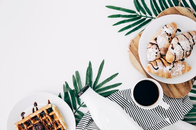 An overhead view of baked croissant; waffles; bottle; coffee cup on leaves over the white backdrop