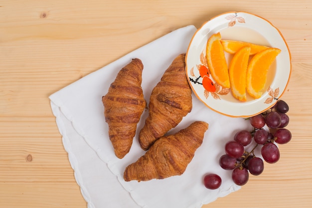 An overhead view of baked croissants; red grapes and orange slices of plate over the wooden desk