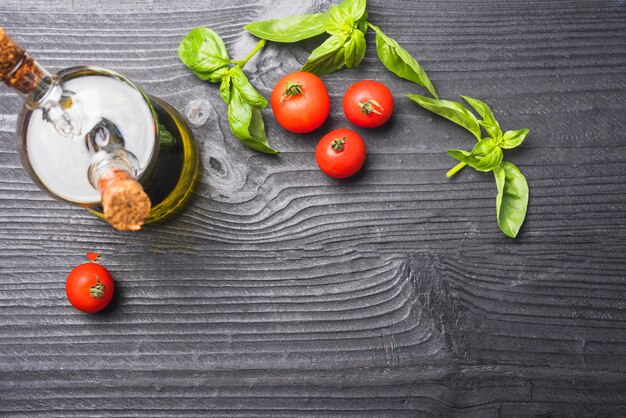 An overhead view of basil leaves; tomatoes and bottle of olive oil on wooden backdrop