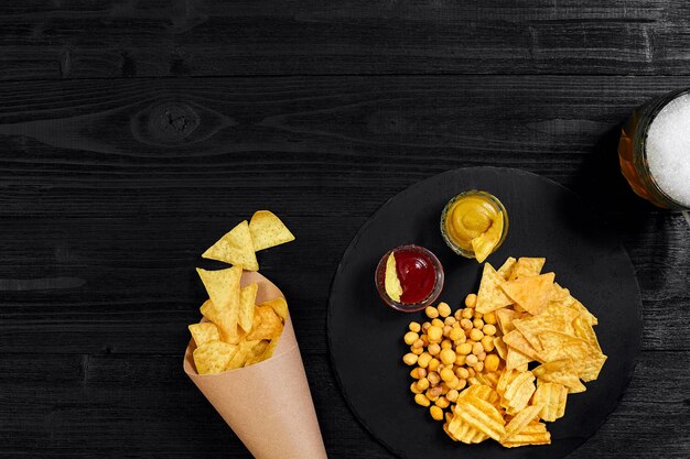 Overhead view of beer glass and snacks with sauce on black wooden table.