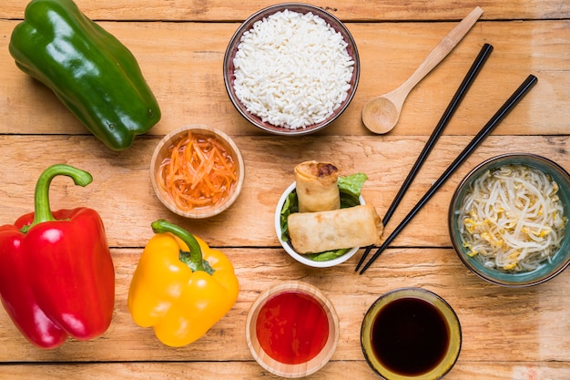 Free photo an overhead view of bell peppers; carrot; rice; spring rolls; sprout beans and sauces with chopsticks and ladle on wooden desk