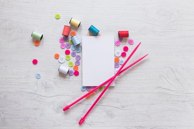 An overhead view of blank paper with spools; button and knitted needle on white background