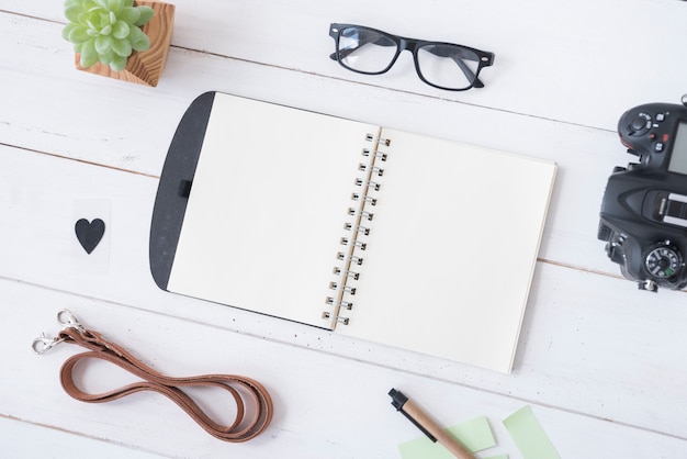 Overhead view of blank spiral notepad; spectacle; belt; pen; heartshape; professional camera and succulent plant on white wooden table