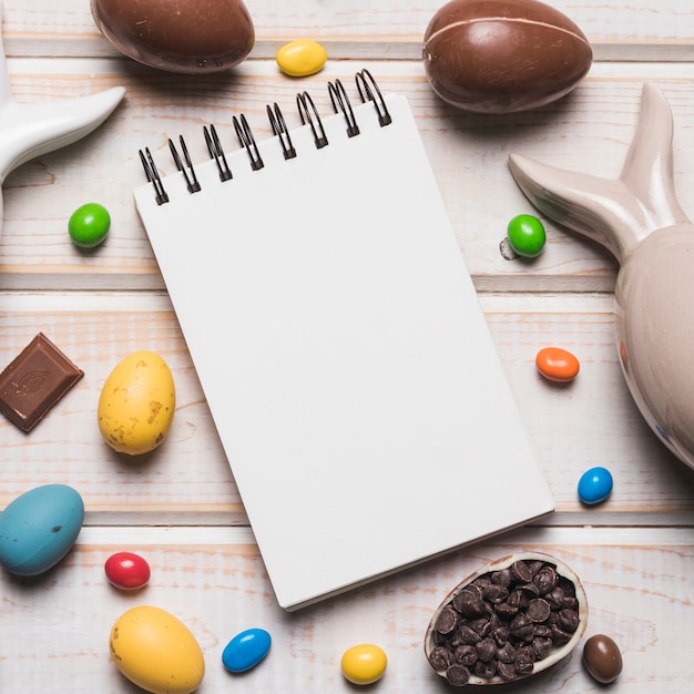 An overhead view of blank spiral notepad with easter eggs; candies and choco chips on wooden desk