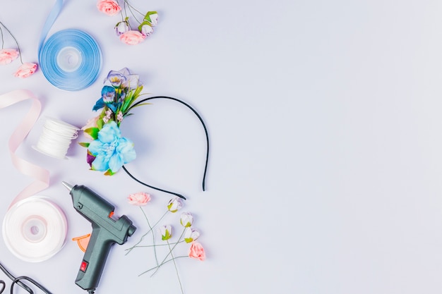 Free photo an overhead view of blue and white ribbon; artificial flower; glue gun for making hairband on white backdrop