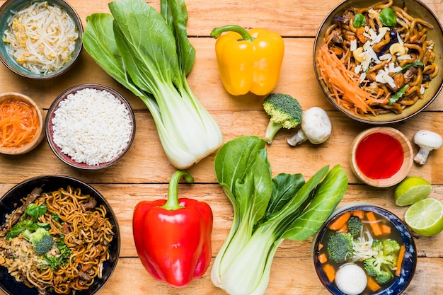 An overhead view of bokchoy; bell peppers; broccoli; mushroom; lemon with thai food on wooden desk