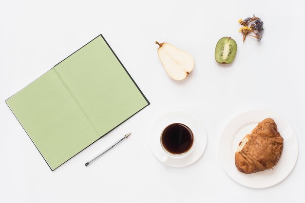 An overhead view of book; pen; halved fruits; coffee and croissant on white background