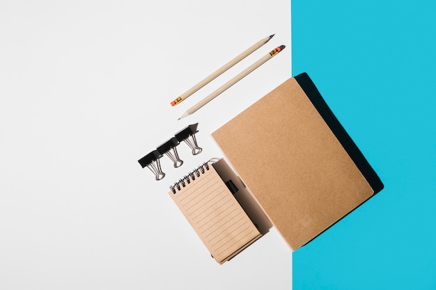 Free photo an overhead view of book; spiral notepad; pencil; bulldog clips on white and blue backdrop