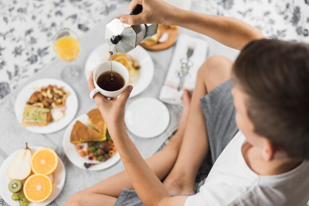 An overhead view of a boy pouring tea in cup with breakfast on table