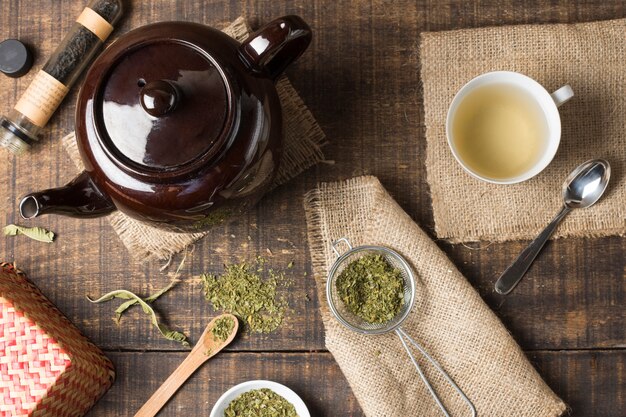 Вид сверху на коричневый чайник; чашка травяного чая и высушенные листья чая на деревянном столе