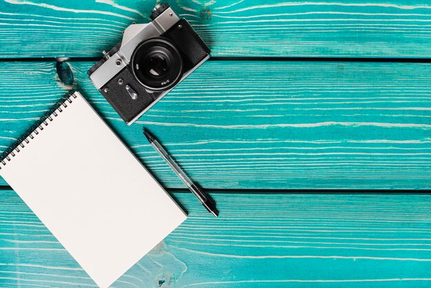 An overhead view of camera; spiral notepad and pen on wooden plank