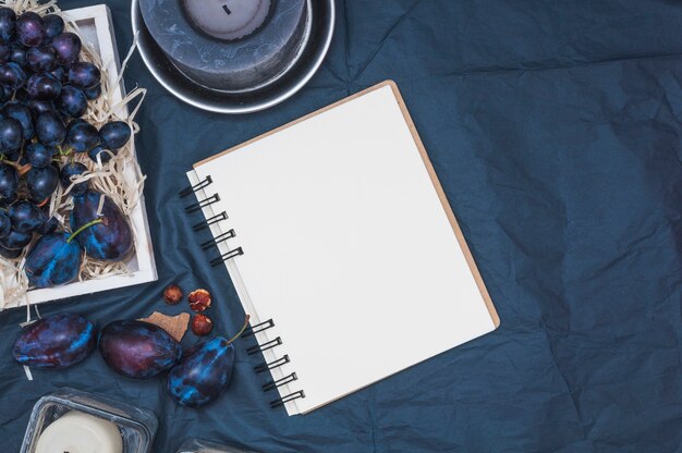 An overhead view of candles; blank spiral notepad; plums and grapes on textile backdrop