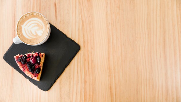 An overhead view of cappuccino coffee with art latte and cake slice on wooden backdrop