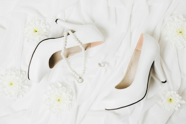 An overhead view of chrysanthemum; necklace; earrings and wedding high heels on white scarf