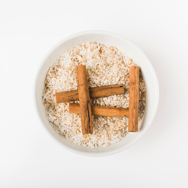 An overhead view of cinnamon sticks with uncooked rice bowl against wooden background