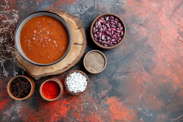 Вид сверху классического томатного супа на деревянном подносе с фасолью и различными специями на столе смешанного цвета