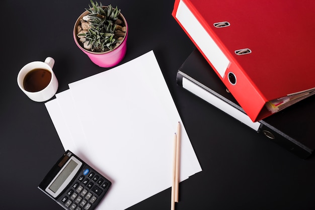 An overhead view of coffee cup; calculator; pot plant; blank white papers; pencils and paper files on black background