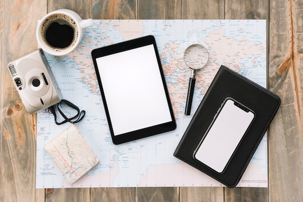 An overhead view of coffee cup; camera; digital tablet; cell phone; magnifying glass and diary on map against wooden table