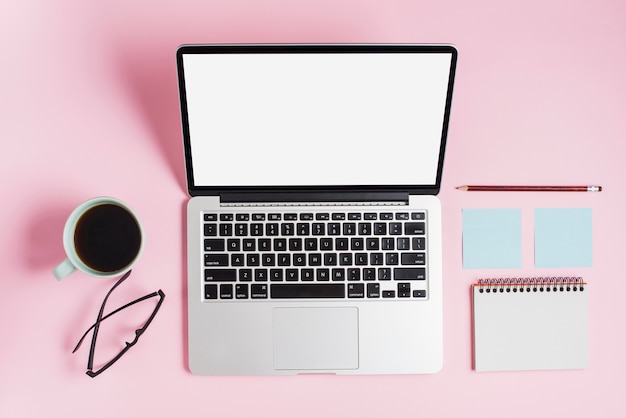 An overhead view of coffee cup; eyeglasses; laptop; pencil; adhesive notepad and spiral notepad against pink backdrop